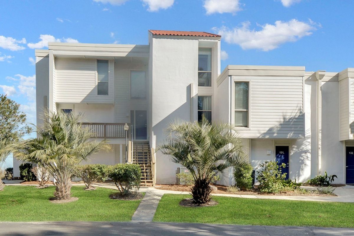 White, modern townhouse building with stairs to the entrance, palm trees, and a green lawn under a blue sky.